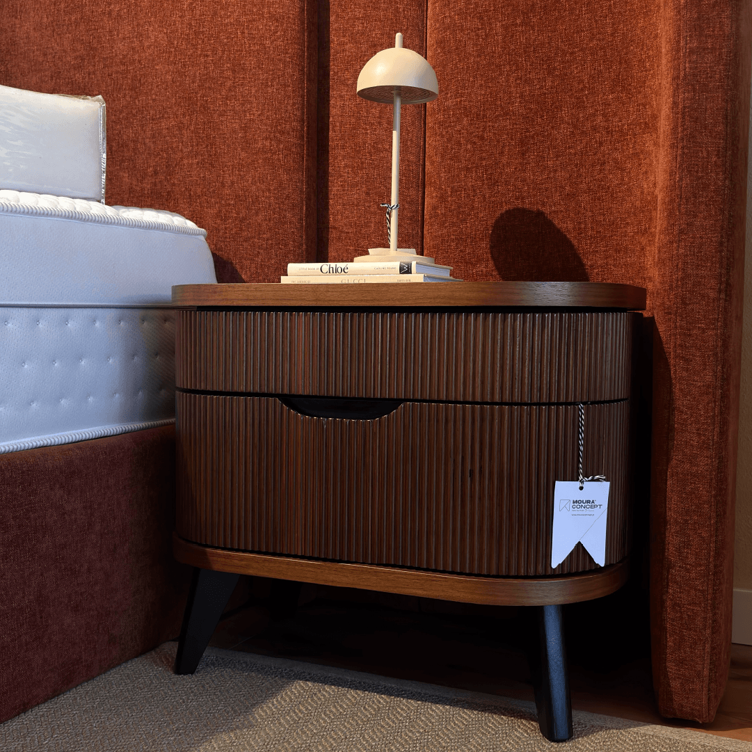 Mesa de cabeceira em madeira ripada com cantos arredondados da Moura Concept, decorada com candeeiro de mesa ao lado de cama com cabeceira estofada a tecido terracota.