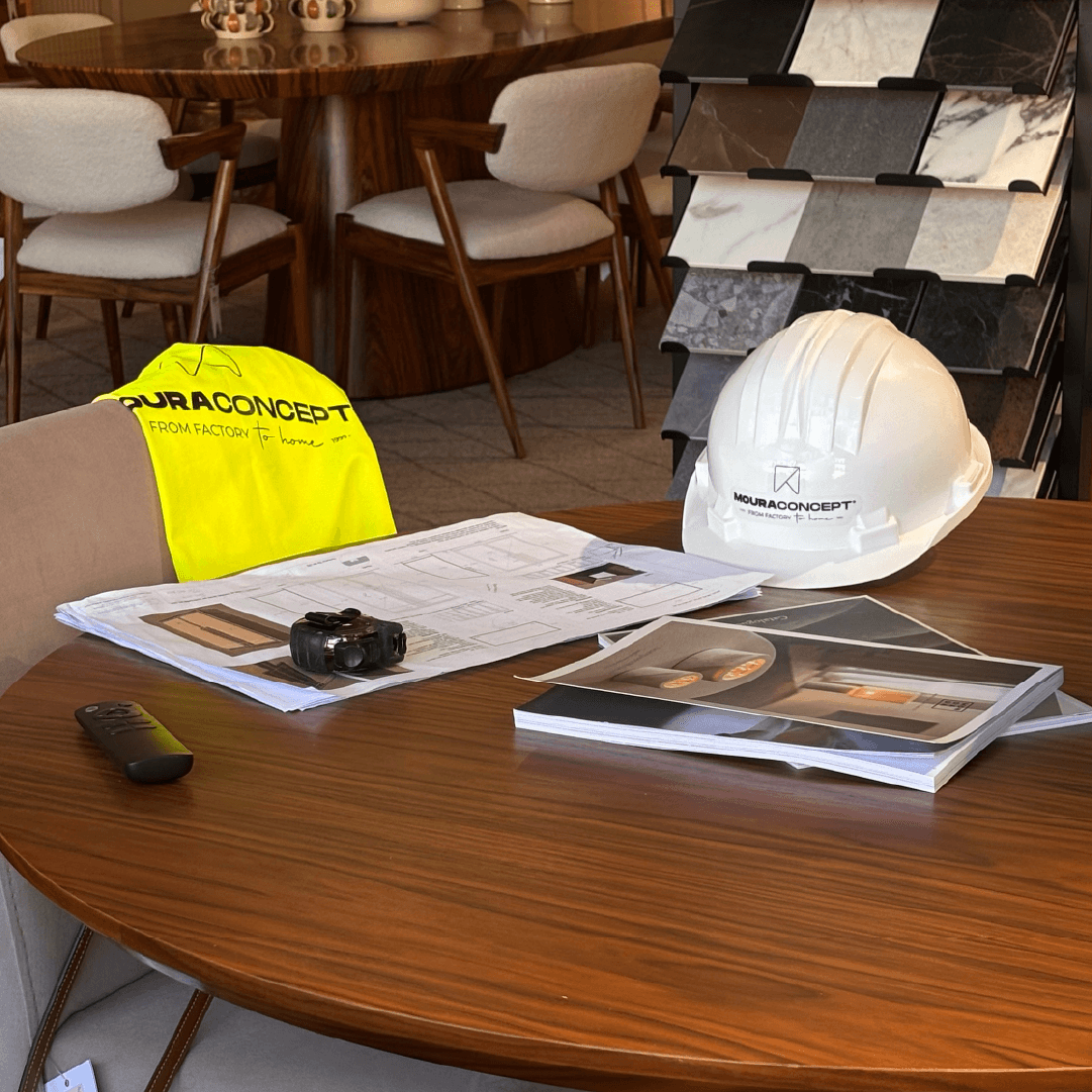 Plantas de arquitetura, capacete e colete corporativos da Moura Concept sobre mesa em madeira, num showroom com mostruário de acabamentos para gestão de obras e projetos chave na mão.